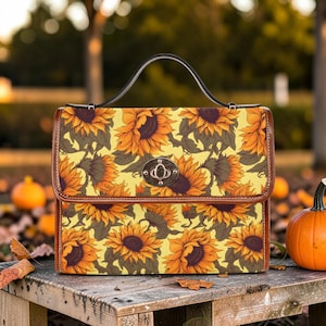 May include: A rectangular handbag with a sunflower pattern. The bag has a brown leather trim, a black handle, and a silver clasp. The background includes pumpkins and autumn foliage.