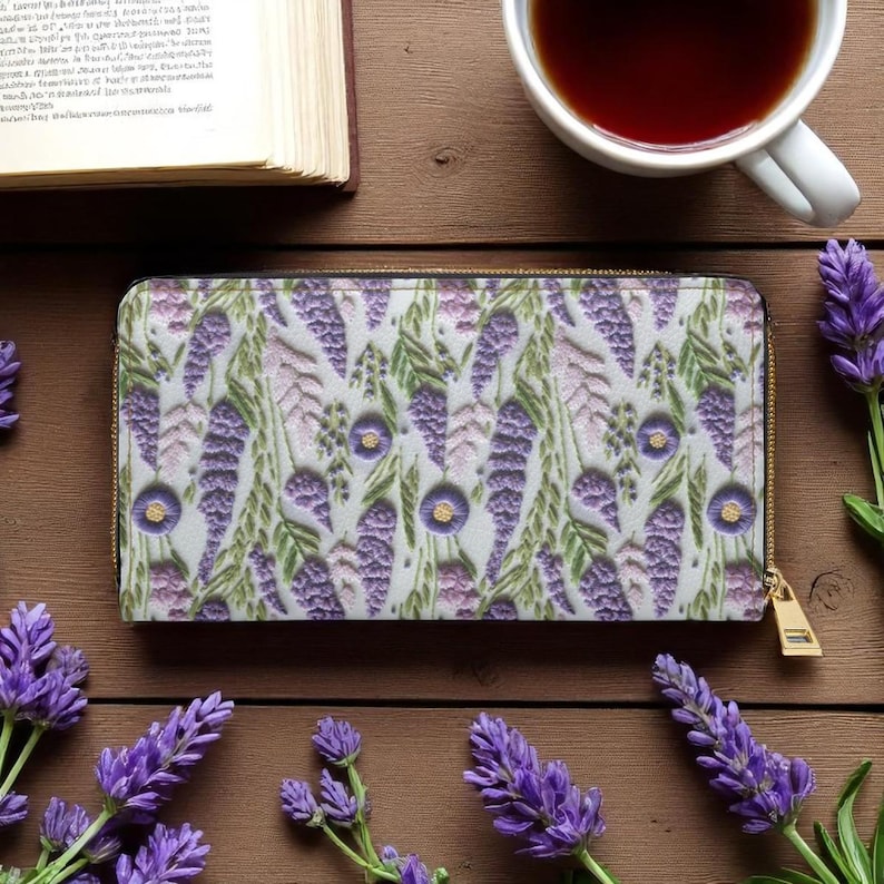 Op de afbeelding: Een rechthoekige portemonnee met een bloemenpatroon van paarse en lavendelkleurige bloemen en groene bladeren. De portemonnee heeft een gouden rits en ligt op een houten oppervlak met een kopje thee en een boek.