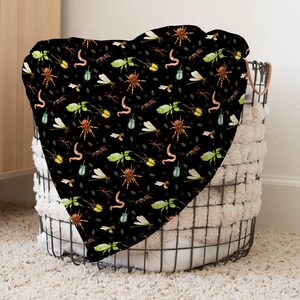 May include: A black blanket with a repeating pattern of insects, including spiders, worms, and dragonflies. The blanket is draped over a wire basket with a fluffy white lining, creating a cozy and whimsical aesthetic.