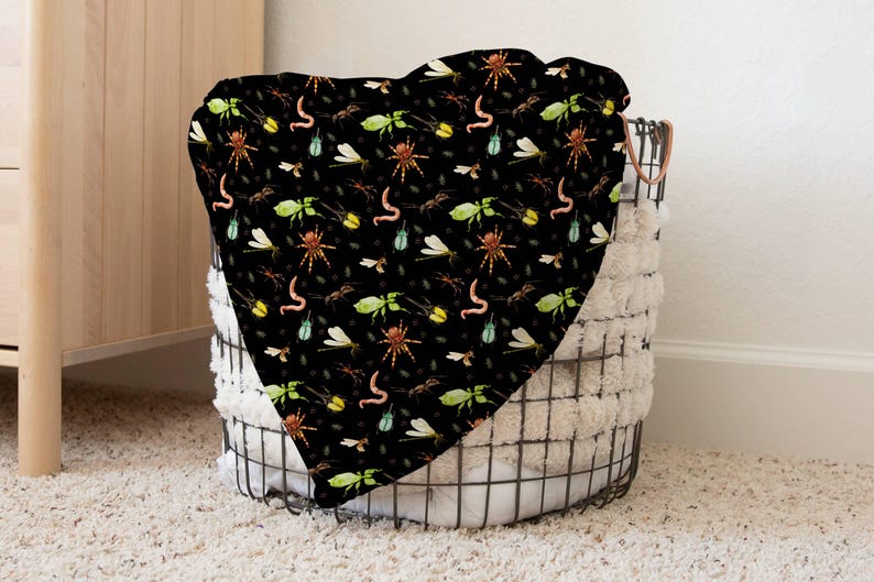 May include: A black blanket with a repeating pattern of insects, including spiders, worms, and dragonflies. The blanket is draped over a wire basket with a fluffy white lining, creating a cozy and whimsical aesthetic.