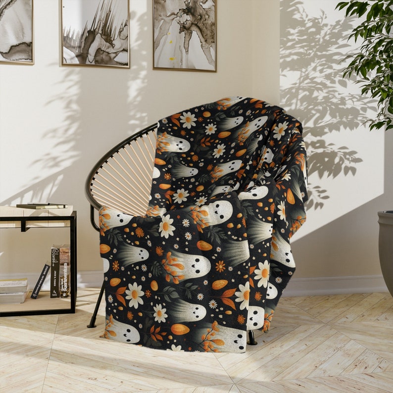 May include: A black and orange throw blanket with a repeating pattern of white ghosts, orange pumpkins, and white flowers. The blanket is draped over a chair.