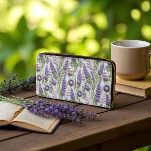 Op de afbeelding: Een witte portemonnee met een bloemmotief van lavendel en groene bladeren. De portemonnee heeft een gouden rits en ligt op een houten tafel. Een kopje koffie en een boek staan ook op de tafel.