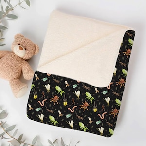 Folded black baby blanket with colorful insects, spiders, and worms, placed next to a teddy bear on a white surface