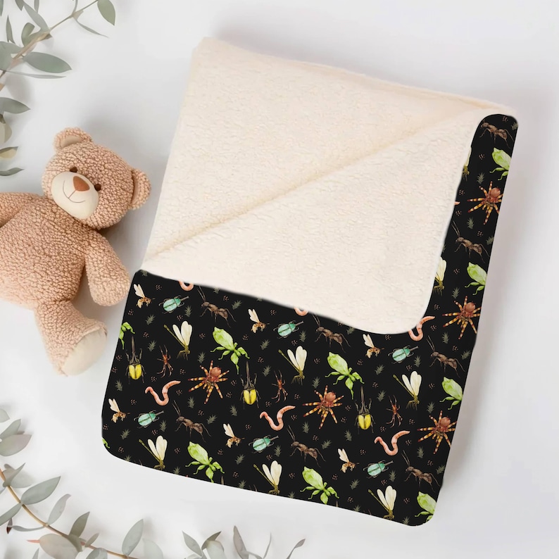 Folded black baby blanket with colorful insects, spiders, and worms, placed next to a teddy bear on a white surface