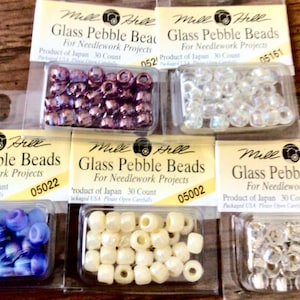May include: Assortment of Mill Hill Glass Pebble Beads for needlework projects. The beads come in various colors, including purple, clear, blue, cream, and silver. Each package contains 30 beads.