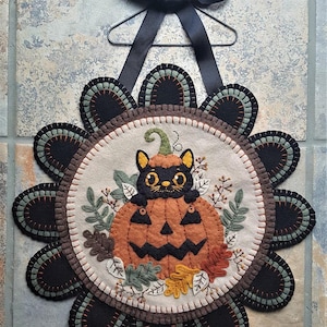 May include: A circular felt wall hanging featuring a black cat sitting inside an orange jack-o'-lantern pumpkin. The design is surrounded by felt leaves and a scalloped border of black and green petals, suspended by a black ribbon and wire hanger.
