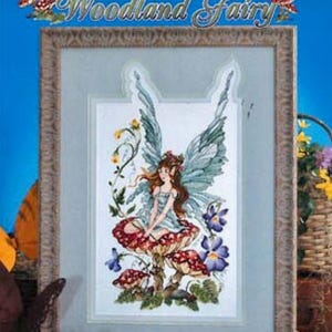 May include: Framed cross-stitch artwork titled "Woodland Fairy" by Stoney Creek Collection. The image features a fairy with blue wings, sitting on a mushroom. The fairy is surrounded by flowers and foliage, with a blue background.