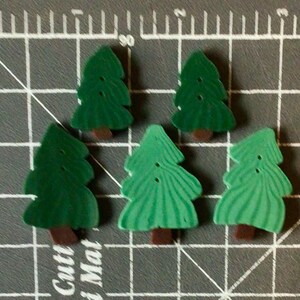 May include: Five green wooden Christmas tree-shaped buttons with brown trunks. The buttons have a textured appearance and small holes for sewing. The trees are arranged on a gray cutting mat.