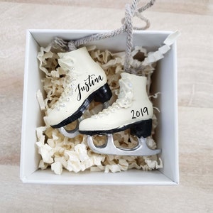 Op de afbeelding: Twee witte ijsschaatsornamenten met zwarte zolen en zilveren messen, gepresenteerd in een witte doos met papieren vulling. Op de ene schaats staat de naam "Justina", op de andere "2019". Een grijze draad is bevestigd om op te hangen.