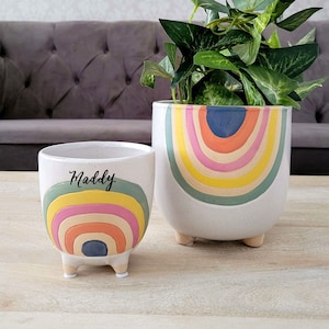 May include: Two rainbow-themed ceramic planters. The larger planter has a white base with a rainbow design and three legs. The smaller planter has a white base with a rainbow design and three legs. The smaller planter has the word "Maddy" written on it.