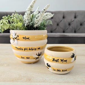 May include: Two ceramic bee-themed planters. The larger planter has white and yellow stripes, with the text "Mom, Thanks for helping me bee me." The smaller planter says "Best Mom in the hive." Both have bee and flower designs.