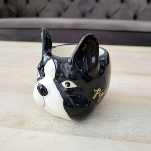 Puede incluir: Una maceta de cerámica con forma de cabeza de perro Boston Terrier. La maceta es negra y blanca con la inscripción dorada "Piper". Las orejas del perro están erguidas y el interior de la maceta es blanco.