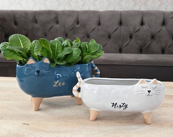 May include: Two cat-shaped ceramic planters, one blue and one white, sit on a wooden table. The blue planter, labeled "Leo", holds green plants. The white planter, labeled "Misty", is empty. Both have wooden legs and cat features.