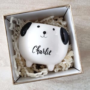 May include: A white ceramic dog-shaped container with black ears and facial features, inscribed with the name "Charlie" in cursive. The container is presented in a gold-edged box filled with shredded paper.