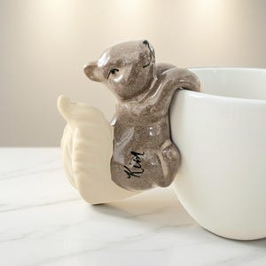 May include: A white ceramic mug with a gray koala bear figurine attached. The koala is hugging the mug, and the word "Kim" is written on the koala's body. The mug is on a white marble surface.
