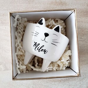 Puede incluir: Una maceta blanca con forma de gato de cerámica con detalles negros, incluyendo orejas, bigotes y nariz. La palabra "Milow" está escrita en cursiva. La maceta está dentro de una caja con borde dorado llena de papel.