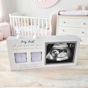 May include: A white rectangular wooden countdown sign featuring two star-shaped toppers, a black and white ultrasound image, and the text Baby Smith, A gift from the stars ready to steal our hearts, and WEEKS UNTIL WE MEET YOU with two flip-style number blocks.