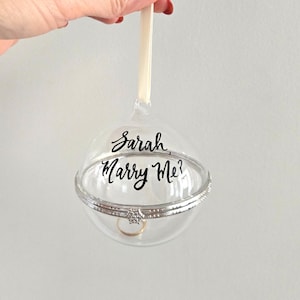 Könnte beinhalten: Ein transparenter Glas-Anhänger mit silbernem Rand und einem Ring im Inneren. Der Anhänger trägt die Aufschrift "Sarah, Marry Me?"