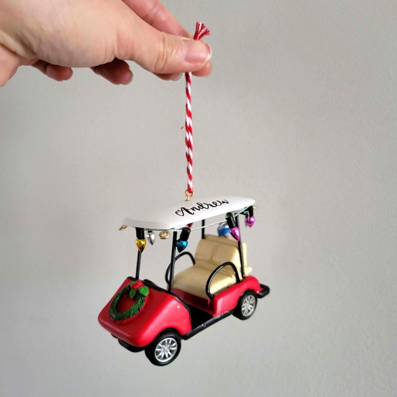 May include: Red golf cart Christmas ornament with a white roof and a wreath on the front. The roof has the name "Andrew" written on it. The ornament has a red and white string for hanging.