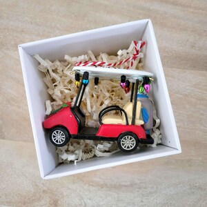 May include: A red golf cart ornament with white trim and a white roof. The cart has a white interior and is decorated with colorful lights. The ornament is sitting in a white box with white shredded paper.
