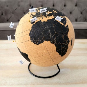 May include: A cork world globe with a black map and a metal stand. The globe has push pins with labels for UK, Italy, Spain, Egypt, China, and Japan.