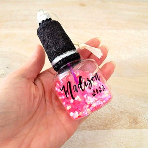 K&ouml;nnte beinhalten: Ein transparenter Glas-Anh&auml;nger in Form einer Nagellackflasche, gef&uuml;llt mit rosa Glitzer. Der Anh&auml;nger hat einen schwarzen Deckel mit einem wei&szlig;en Streifen und den Namen "Madison" sowie das Jahr "2022" darauf geschrieben.