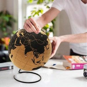 May include: A cork globe with a black world map and a metal stand. The globe is being used to mark locations with push pins.