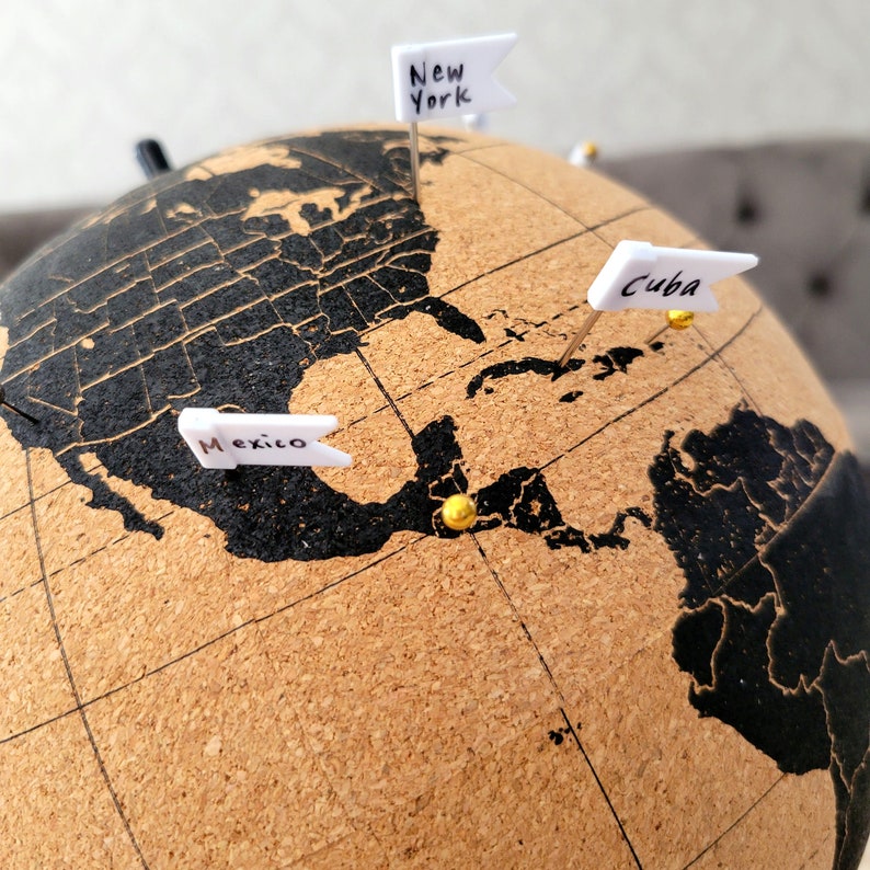 May include: A corkboard globe with black continents and gold push pins. The globe has flags with the words "New York", "Mexico", and "Cuba" on them.