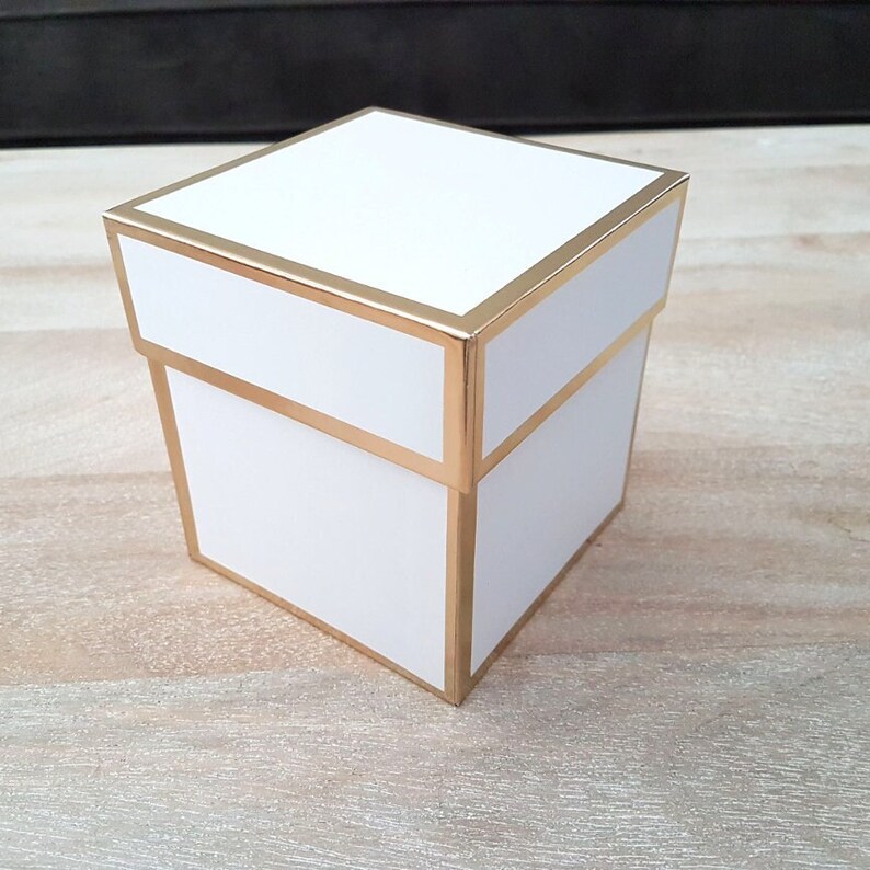 May include: A white square gift box with a gold trim around the edges. The box is sitting on a light wood surface.