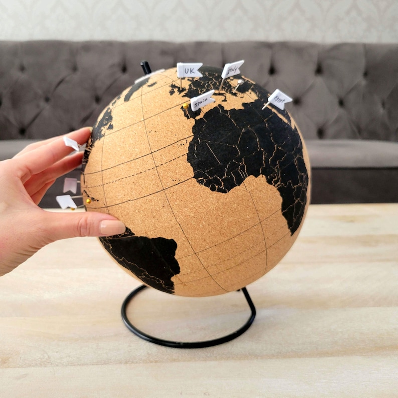 May include: A cork globe with a black map and a metal stand. The globe has push pins with white flags that have handwritten locations on them, including "UK" and "Spain".