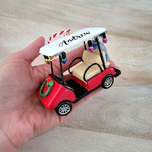May include: Red golf cart Christmas ornament with a white roof and the name "Andrew" written on it. The cart has a wreath on the side and is decorated with colorful lights.