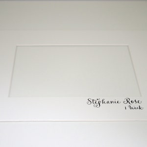 Puede incluir: Una cartulina blanca con una abertura rectangular y el texto "Stephanie Rose 1 Week" en escritura cursiva negra.