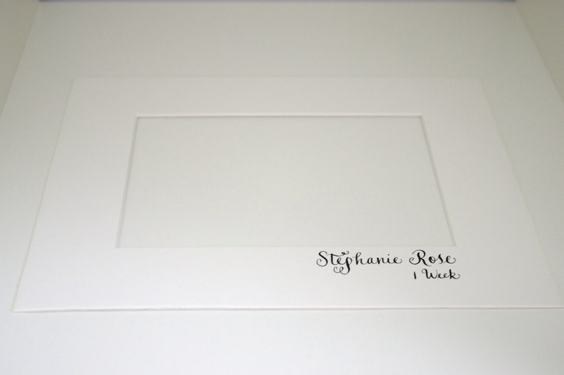Puede incluir: Una cartulina blanca con una abertura rectangular y el texto "Stephanie Rose 1 Week" en escritura cursiva negra.