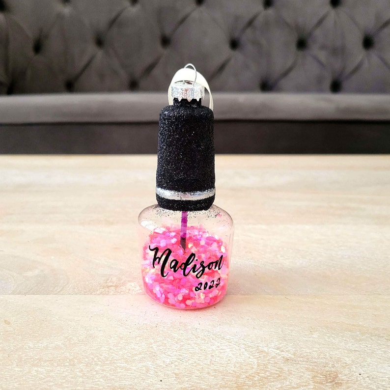 K&ouml;nnte beinhalten: Ein durchsichtiger Nagellack-Flaschen-Anh&auml;nger aus Glas mit einem schwarzen Glitzer-Deckel und einer rosa Glitzer-Basis. Die Flasche tr&auml;gt den Namen "Madison" und das Jahr "2022" ist darauf geschrieben.