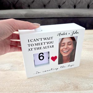 Puede incluir: Un bloque de cuenta regresiva blanco con el texto "I CAN'T WAIT TO MEET YOU AT THE ALTAR" y una foto de una persona sonriente. Se muestra el número "6", junto con un corazón rojo. Los nombres "Amber & John" están escritos encima de la foto.