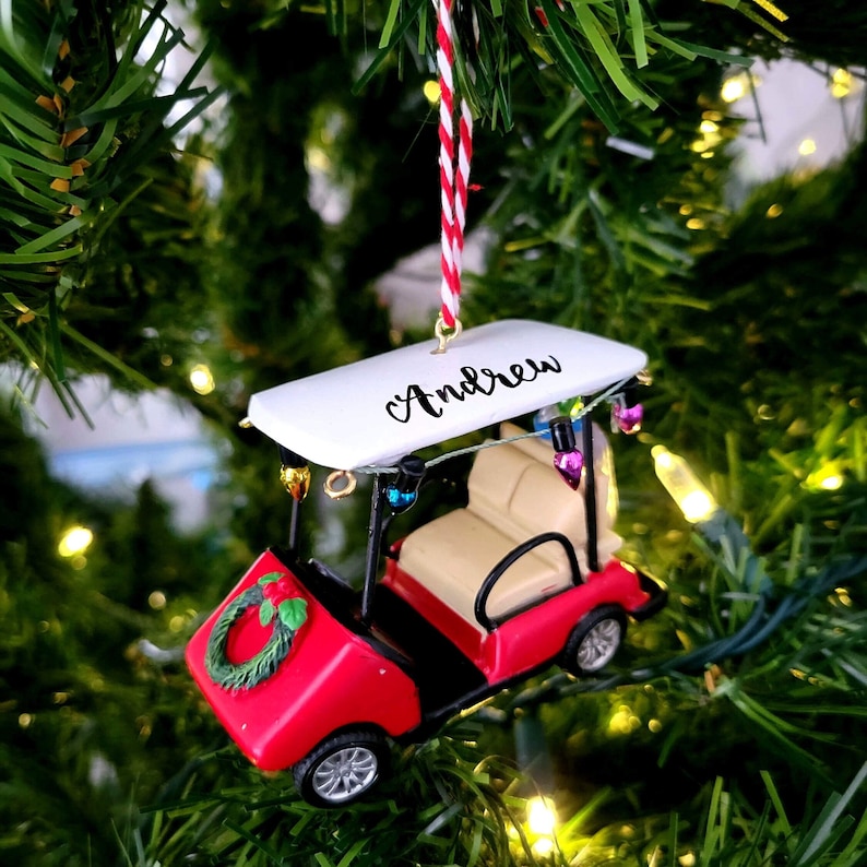 May include: Red golf cart Christmas ornament with a white roof that says "Andrew" in black lettering. The cart has a wreath on the front and lights strung around the top.