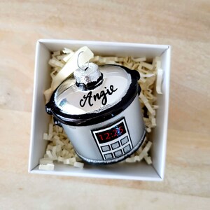 May include: A silver and black glass ornament shaped like a slow cooker with a digital timer display. The ornament is personalized with the name "Angie" on the top.