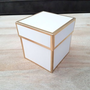 Op de afbeelding: Een witte cadeaudoos met een gouden rand. De doos is vierkant en heeft een deksel.