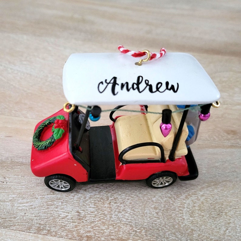 May include: Red golf cart Christmas ornament with a white roof, a green wreath, and a string of lights. The roof is personalized with the name "Andrew".