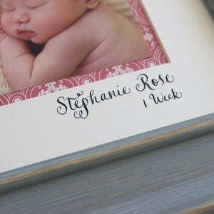 Puede incluir: Un marco de fotos blanco con un borde de patr&oacute;n rosa y blanco. El marco contiene una foto de una ni&ntilde;a beb&eacute; con el texto "Stephanie Rose 1 Semana" escrito debajo de la foto.
