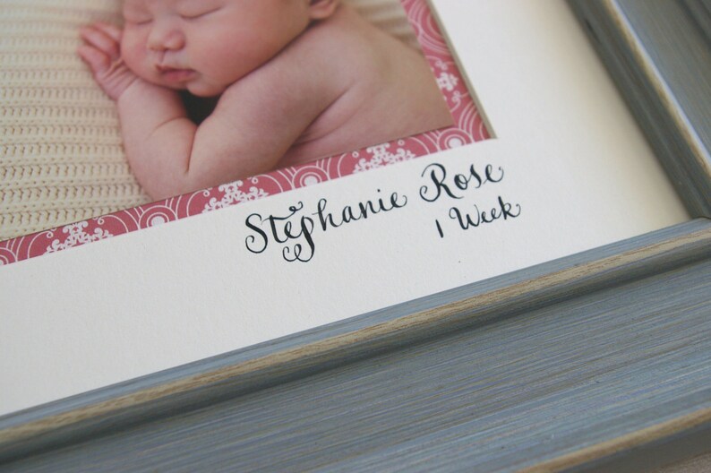 Puede incluir: Un marco de fotos blanco con un borde de patr&oacute;n rosa y blanco. El marco contiene una foto de una ni&ntilde;a beb&eacute; con el texto "Stephanie Rose 1 Semana" escrito debajo de la foto.