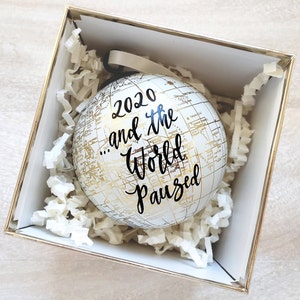 Puede incluir: Un adorno navideño de globo terráqueo blanco con detalles dorados y el texto "2020...and the world paused" en caligrafía negra.