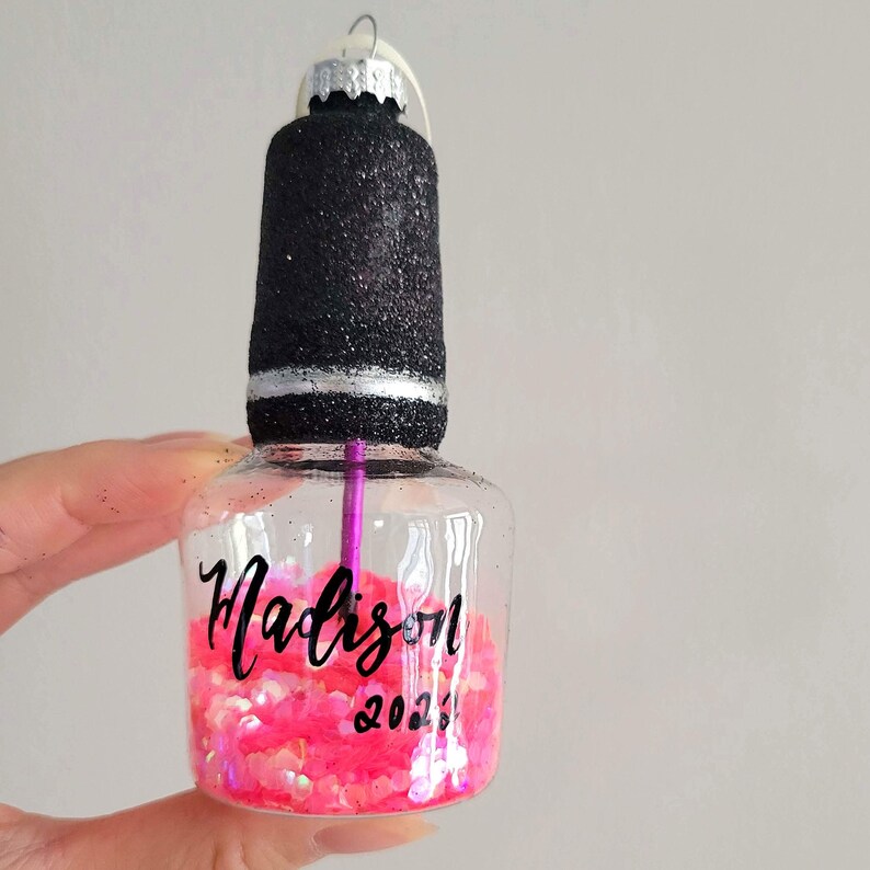 K&ouml;nnte beinhalten: Eine durchsichtige Glasflasche in Form eines Nagellackflaschens mit einem schwarzen Glitzerdeckel und einem silbernen Band. Die Flasche ist mit rosa Glitzer gef&uuml;llt und tr&auml;gt den Namen "Madison" und das Jahr "2023" darauf.