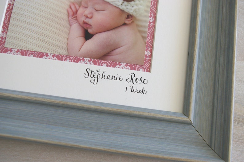 Puede incluir: Un marco de fotos blanco con un borde de madera azul claro. El marco contiene una foto de un beb&eacute; durmiendo con un borde estampado en rosa y blanco. El texto "Stephanie Rose 1 Week" est&aacute; escrito debajo de la foto.