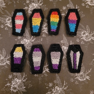 May include: Eight embroidered coffin-shaped patches with different pride flag designs. The flags include transgender, bisexual, pansexual, lesbian, genderqueer, asexual, and non-binary.