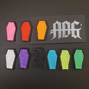 May include: A collection of embroidered coffin patches in various colors, including pink, red, orange, white, purple, black, blue, lime green, and green. A black rectangle with the text "ADG" in a gothic font is also present.