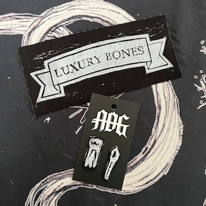 May include: A black and white patch with the text "Luxury Bones" on a black background. A black card with the text "AOG" and two white embroidered patches of a tooth and a bone.