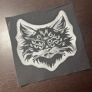 Könnte beinhalten: Weiß gestickter Katzenkopf-Patch mit mehreren Augen auf schwarzem Stoffhintergrund.