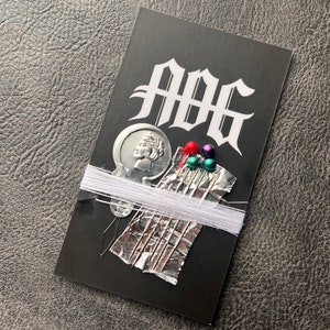 May include: A black card with the word "ABB" printed in white. The card has a silver coin with a profile of a person on it, white thread, and a small piece of aluminum foil with several sewing needles and colorful beads.