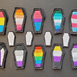 May include: A set of 12 embroidered patches shaped like coffins, each featuring a different pride flag design. The flags include the pansexual, transgender, lesbian, bisexual, genderqueer, non-binary, asexual, intersex, and rainbow pride flags.
